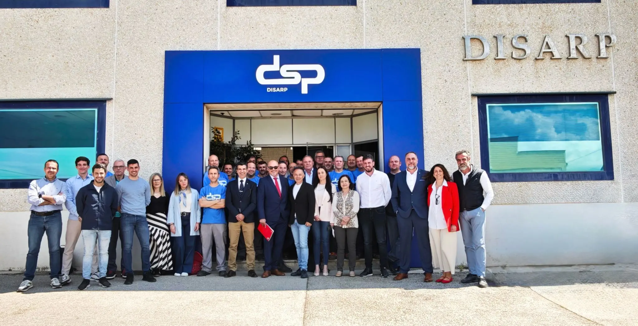 Fotografía del equipo humano de DSP posando frente a la sede principal, junto a la entrada con el logotipo azul corporativo.