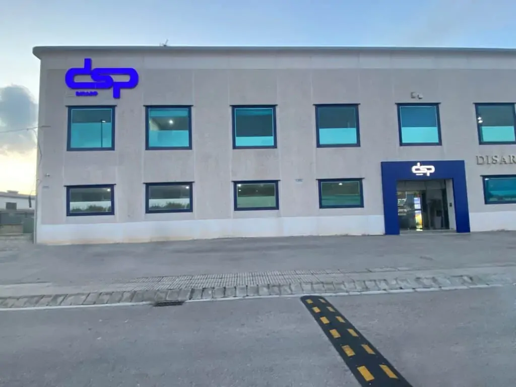 Vista frontal del edificio principal de DSP con fachada clara, ventanas azules y el logotipo corporativo visible en el acceso principal y en la parte superior izquierda del edificio.