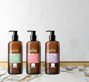 Tres botellas de productos ecológicos MIORI sobre superficie de mármol: acondicionador capilar (verde), champú y acondicionador 2 en 1 (rosa), y gel de ducha y champú (lila), todos con certificación EU Ecolabel.